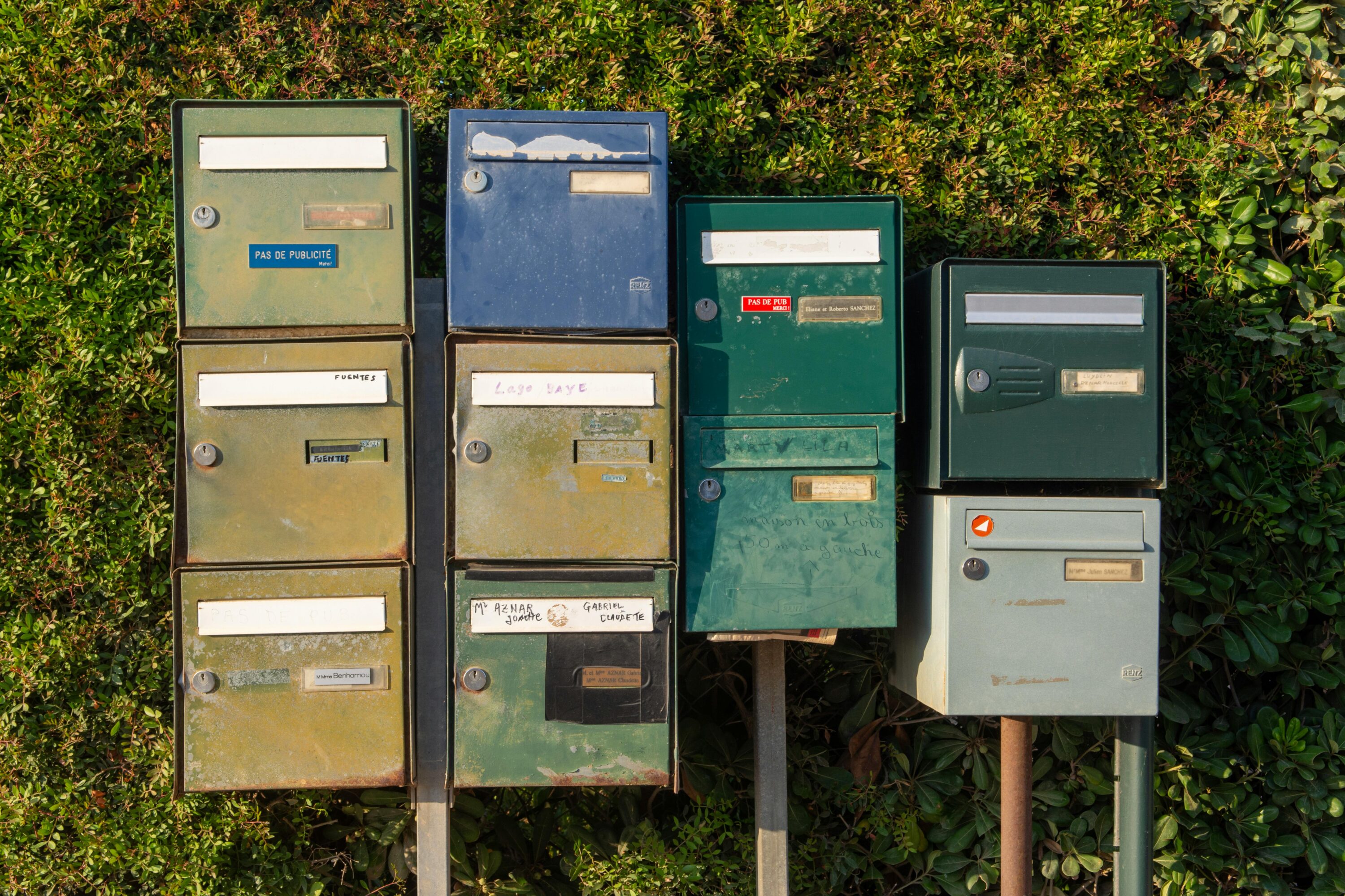 A collection of vintage metal mailboxes on posts against a hedge backdrop.